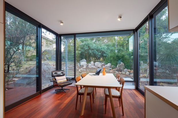 A bright, glass-enclosed dining room or sunroom with floor-to-ceiling glazing on three sides, a rectangular dining table with timber chairs, and a mid-century-style lounge chair. Dark-framed windows overlook a landscaped rocky garden, while rich reddish timber flooring and a white ceiling create a warm, airy interior.