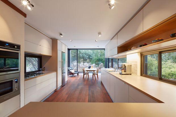 A long, contemporary galley kitchen opening into a dining and living area. The space features white flat-panel cabinetry, integrated stainless-steel appliances, a light benchtop, warm timber flooring, and a timber-lined open display shelf above the right-hand counter. Large windows and full-height glazing provide views into dense greenery and bring natural light into the room.