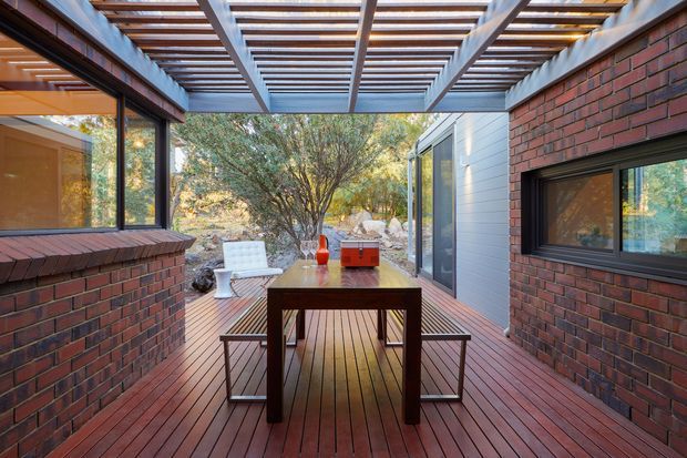 A covered outdoor dining terrace enclosed by red brick walls and large dark-framed windows, with a timber deck, timber pergola, and exposed steel beams. A wooden dining table and chairs occupy the centre, while a light-clad glazed extension and landscaped bush setting are visible beyond.