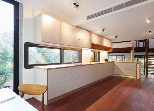 A bright open-plan kitchen with extensive white built-in cabinetry, warm timber upper cabinet fronts, a long pale benchtop, and a horizontal splashback window overlooking greenery. Dark timber flooring, black-framed glazing, integrated linear air-conditioning vents, and a stairway/mezzanine area are visible. The space has a clean, warm modern character with abundant natural light.