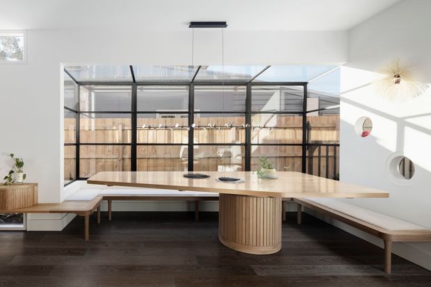 Bright contemporary dining area with a long pale timber dining table, integrated curved pedestal base, and built-in bench seating wrapping around the room. A large black-framed glazed enclosure opens the space to a fenced outdoor area, while white walls, dark timber flooring, sculptural pendant lighting, and circular wall mirrors create a clean, refined interior.