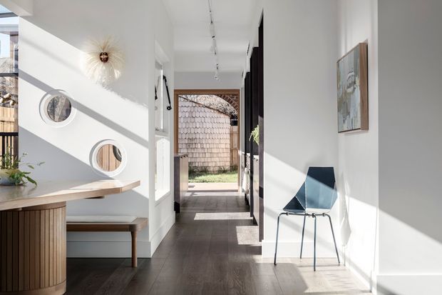A bright, contemporary residential hallway or entry passage with white walls, dark timber flooring, and strong diagonal sunlight. On the left is a built-in timber dining or breakfast nook with a bench and circular mirrors, while a minimalist dark-framed chair and artwork sit along the right wall. The corridor leads toward an exterior courtyard or garden, with timber shingle cladding visible beyond.