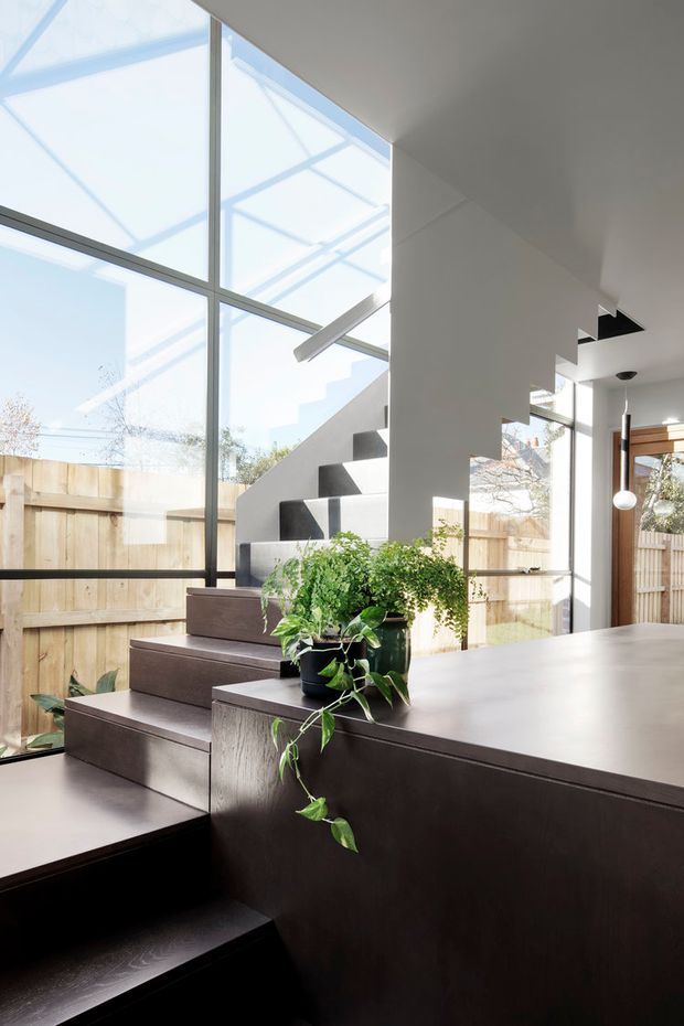 A bright contemporary interior centered on a sculptural staircase beside a dining or kitchen island. Floor-to-ceiling glazing brings in abundant daylight and reveals a timber boundary fence outside. The staircase has dark timber treads with a white geometric side wall, while the large dark timber built-in island or joinery unit forms the foreground. Indoor plants and a slender pendant light add natural and refined accents.