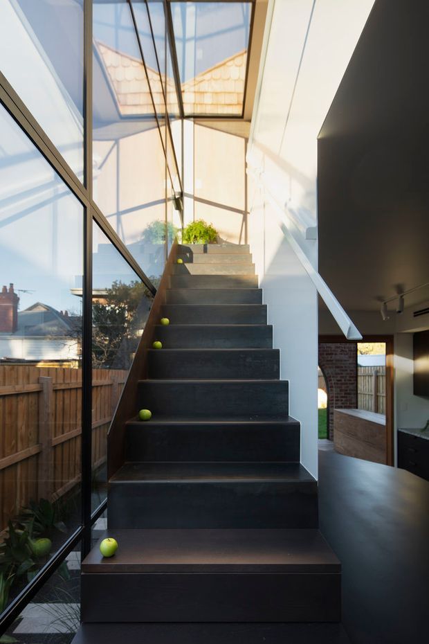 A dramatic interior stairway with dark, solid-looking stair treads and risers ascending beside full-height glazed windows. The stair rises toward a bright landing with a potted plant, while exposed brick and dark cabinetry or wall surfaces are visible to the side. Strong daylight, structural glazing, and crisp geometric lines create a modern, sculptural atmosphere.