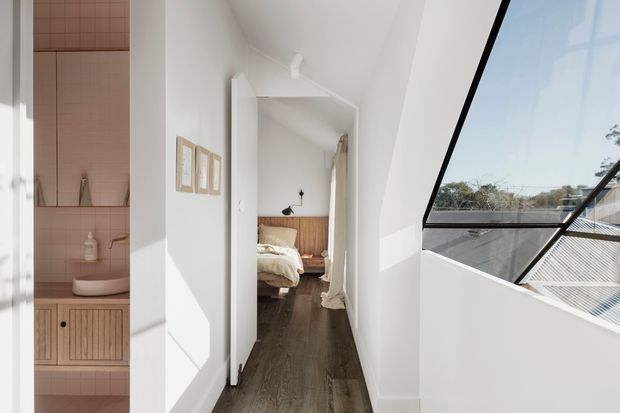 A bright, narrow residential hallway viewed from inside, with a compact pink-tiled bathroom and wall-mounted basin on the left, a bedroom visible through an open doorway at the end, and a large angled glazed opening on the right. The interior has white walls, dark timber-look flooring, and strong natural light.