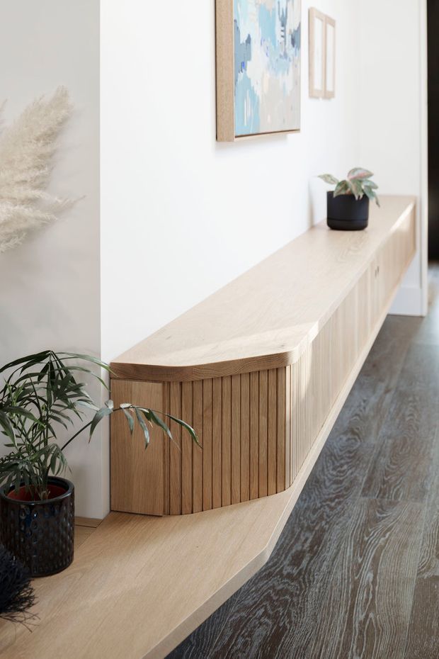 A bright interior hallway or entry passage featuring a long, built-in light timber console and bench with curved ends and vertical slatted cabinetry. White walls display abstract artwork, while potted plants add greenery. Dark timber flooring contrasts with the pale wood joinery, creating a calm, refined contemporary setting.