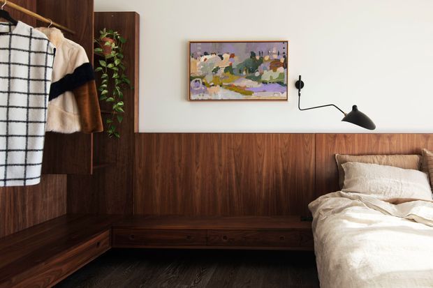 A warm, refined bedroom featuring extensive dark timber wall panelling and integrated built-in storage, including a low bench or drawer unit. A partially visible bed with rumpled cream bedding occupies the right side, while a dark sculptural wall-mounted reading lamp and colourful framed artwork provide accents. Clothing hangs at the left beside a tall timber wardrobe, and a leafy plant adds a small natural element.