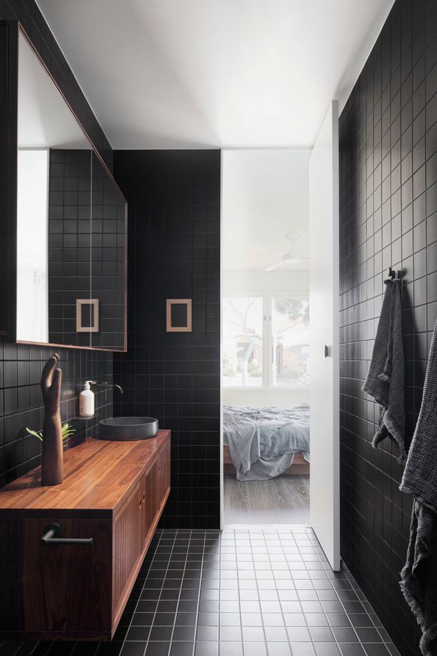 A contemporary bathroom viewed toward an open doorway into a bedroom. The bathroom features full-height matte black square wall tiles, matching dark floor tiles, a floating dark timber vanity with a vessel basin and wall-mounted tap, a large frameless mirror, and dark towels on wall hooks. Bright natural light enters from the bedroom beyond, creating strong contrast between the dark bathroom and the lighter adjoining space.