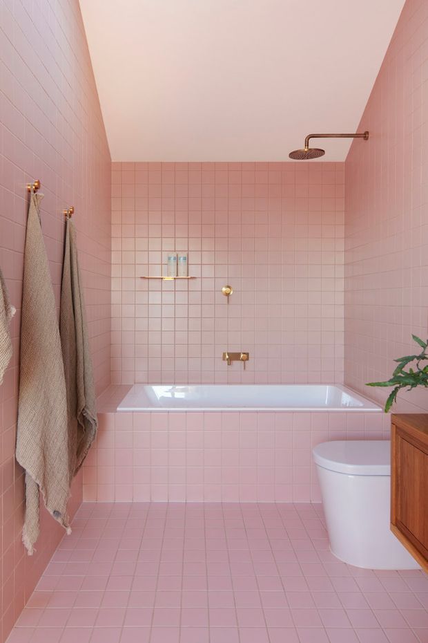 A compact pink-tiled bathroom with a built-in white bathtub, wall-mounted rainfall shower, white toilet, and timber vanity cabinetry. Brass-gold plumbing fixtures and towel hooks add warmth, while neutral textured towels and a small plant provide restrained decoration. The room has a simple rectangular layout, tiled walls and floor, and a clean, playful monochromatic palette.