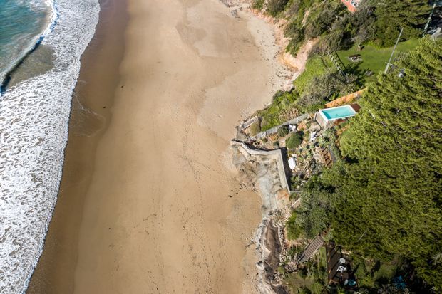 Aerial view of a sandy beach and surf beside a densely vegetated coastal property. A small turquoise swimming pool, terraces, paths, and retaining structures are visible along the shoreline edge.