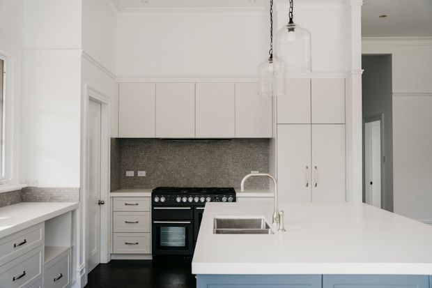 Bright, refined kitchen featuring white built-in cabinetry, a large pale island with integrated sink, dark freestanding range, grey mosaic tile splashback, dark flooring, pendant glass lights, and traditional wall mouldings.