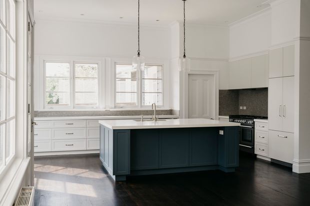 Bright, spacious kitchen with a large dark navy island, white perimeter cabinetry, a pale stone benchtop, dark timber flooring, and a textured dark splashback. Tall windows provide abundant natural light, while decorative wall panelling, cornices, pendant lights, and traditional detailing create a refined classic character.