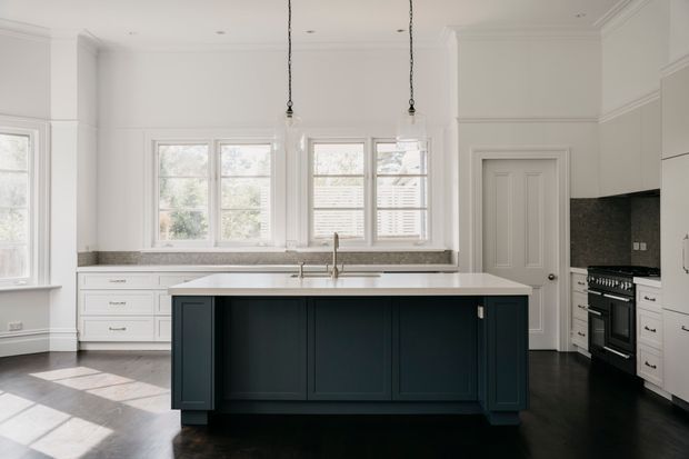 Bright, refined traditional kitchen with a large dark navy island, white Shaker-style cabinetry, pale stone benchtops, dark timber flooring, tall sash windows with shutters, and classic wall and ceiling mouldings. Pendant lights hang above the island, while a range cooker and dark splashback sit along the right wall.