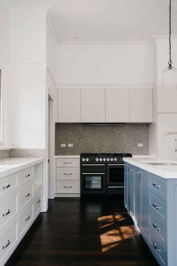 Bright, traditional-style kitchen with a compact galley layout. White perimeter cabinetry and upper cupboards frame a black freestanding range, while a pale blue island with shaker-style drawers occupies the right side. The kitchen features a small grey mosaic tile splashback, dark timber flooring, white stone-look benchtops, high decorative ceiling cornices, and a pendant light.