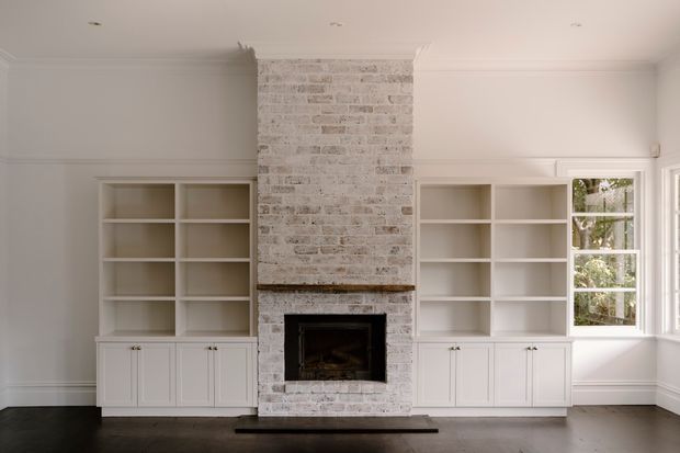 Bright, refined living room featuring a full-height white-painted brick fireplace with a dark firebox, symmetrical built-in white shelving and lower cabinetry, wall moulding, and a projecting bay window. Dark timber flooring provides contrast against the predominantly white interior.