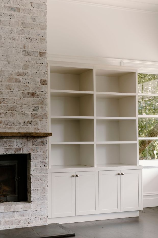 A bright, refined living room interior featuring a white-painted brick fireplace with a dark timber mantel, bespoke white built-in shelving and lower cupboards, decorative wall moulding, a large divided window, and grey tiled flooring.