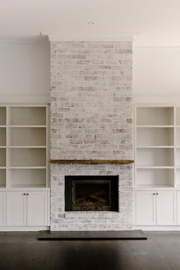 A refined living room feature wall with a tall whitewashed exposed-brick fireplace, dark timber mantel shelf, and black firebox. Symmetrical built-in white shelving and closed cabinetry flank the fireplace, with decorative ceiling cornices and dark timber flooring. The restrained palette and traditional detailing create a calm, understated interior.