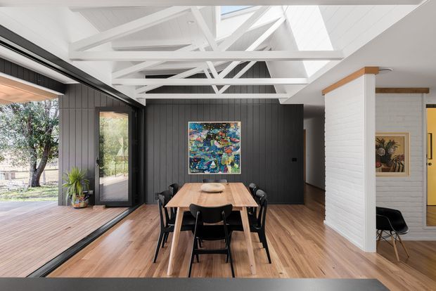 Bright contemporary dining area with a timber dining table and black chairs, positioned beside expansive glazed sliding doors opening to a timber deck. The room features warm timber flooring, dark vertical wall panelling, white painted exposed roof trusses and ceiling boards, skylights, and a white brick-lined partition. Colourful artwork and indoor plants add accents.