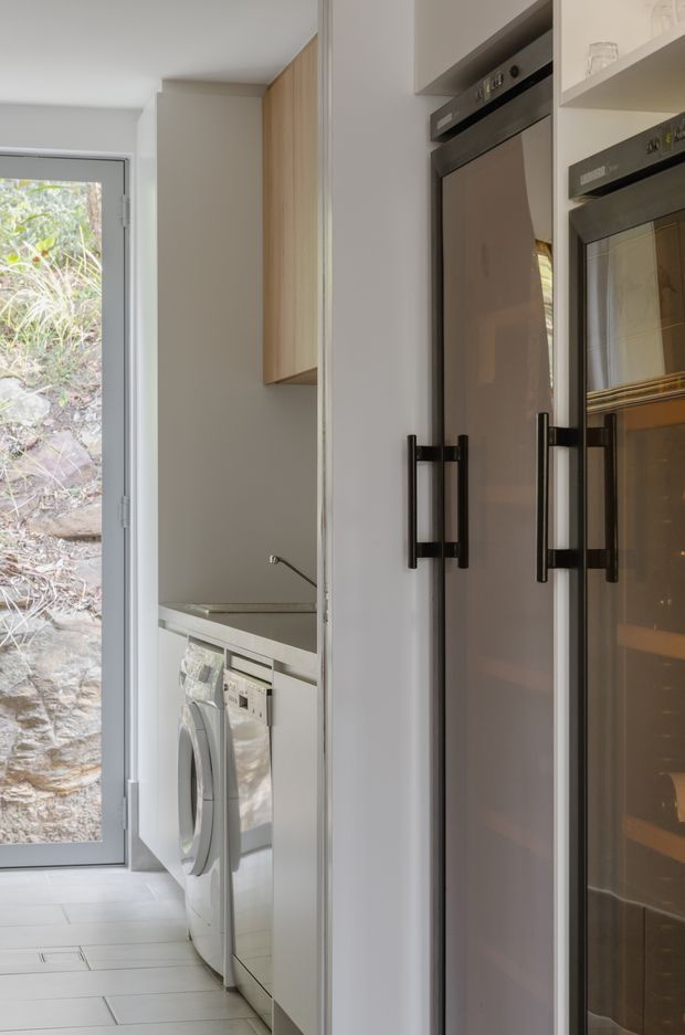 A narrow, bright utility area with a front-loading washing machine beneath a pale benchtop, light wood overhead cabinetry, and tall dark-framed built-in appliances, likely ovens or refrigeration. A full-height glazed exterior door brings in natural light and reveals a stone retaining wall outside. The space has a clean, minimalist built-in fit-out in neutral tones.