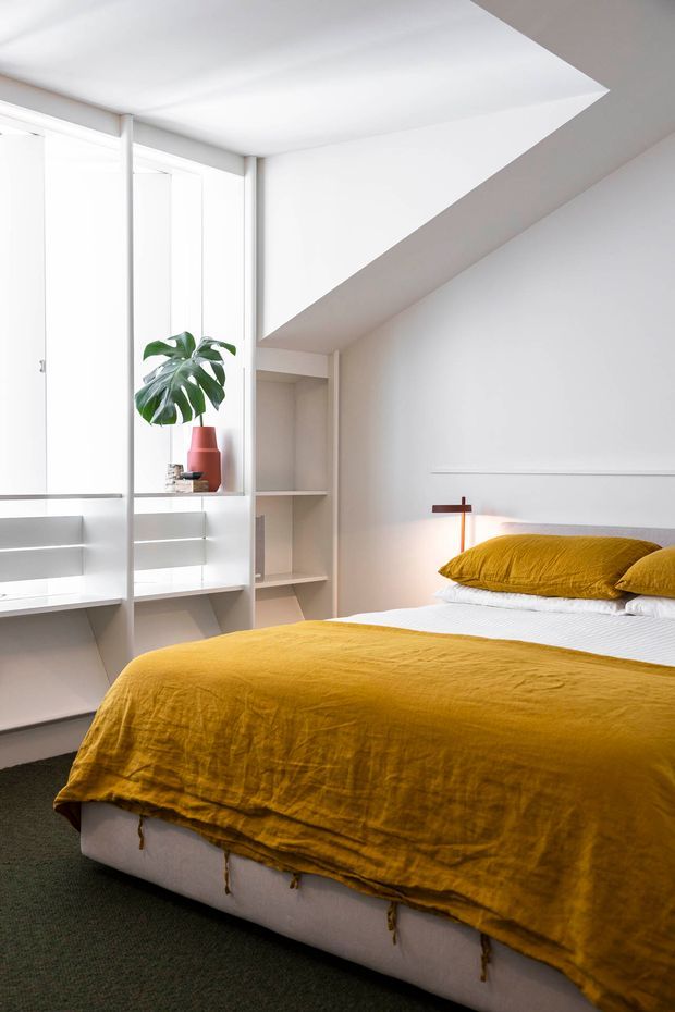 A bright, contemporary bedroom with a low-profile bed dressed in mustard-yellow linen bedding. White built-in shelving and cabinetry frame tall windows on the left, with a potted monstera and small decorative objects. A simple wall-mounted bedside light and geometric angled ceiling detail add architectural interest. The room has white walls and joinery, dark carpet, and a clean, uncluttered composition.