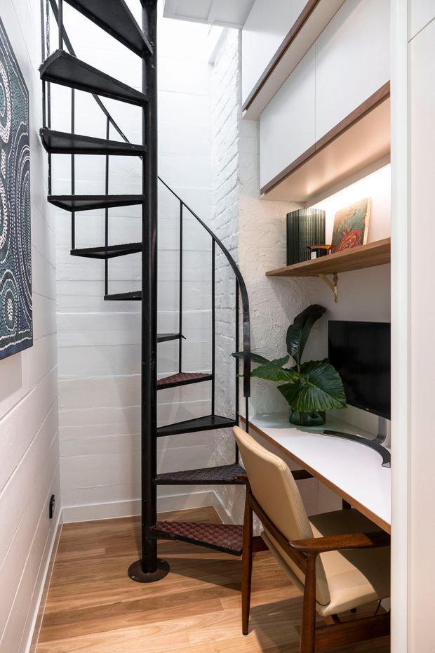 Compact home office nook beside a striking black metal spiral staircase. The space features white painted masonry walls, warm timber flooring, a built-in white desk and cabinetry with timber shelving, integrated warm lighting, a mid-century-style leather-and-wood chair, artwork, and a potted plant.