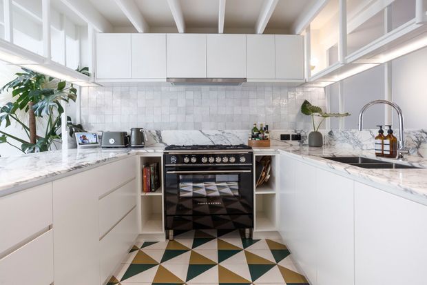 Bright contemporary kitchen with white flat-panel cabinetry, marble-look benchtops, a white textured tiled splashback, and a freestanding black range cooker. The room features exposed white ceiling beams, extensive glazing, a patterned geometric tiled floor, and indoor plants. A large island or peninsula with an undermount sink and gooseneck tap occupies the foreground.