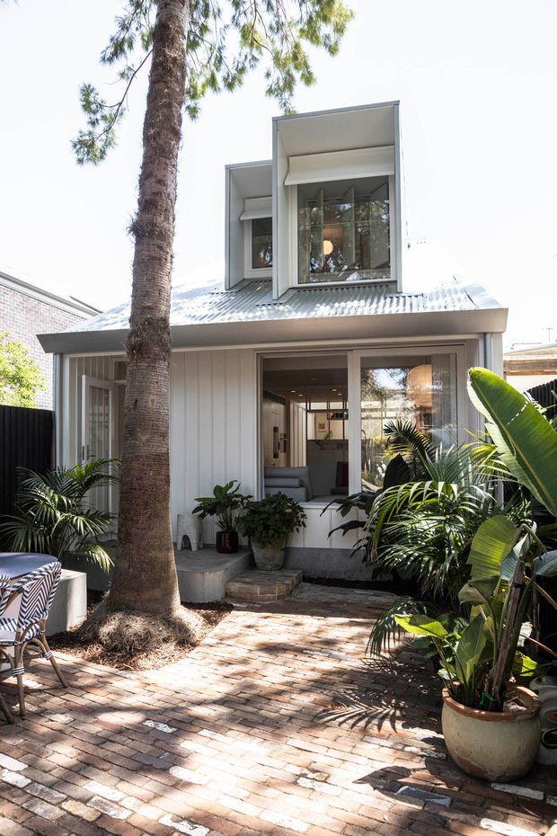 Bright exterior view of a compact two-storey residence from a brick-paved courtyard. The house has a white vertical-panel facade, corrugated metal roof, projecting upper-level box windows with deep reveals, and large glazed openings revealing a living area inside. The courtyard is densely planted with tropical foliage and includes outdoor seating.