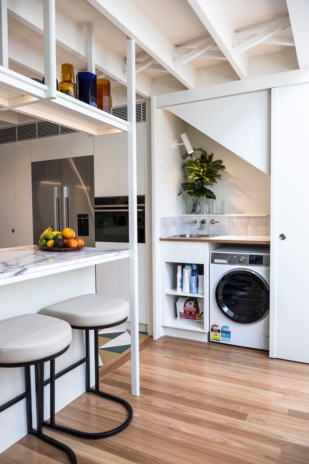 Bright, compact open-plan kitchen with an integrated laundry nook. White structural ceiling beams and shelving frame stainless-steel appliances, a marble-look island benchtop, bar stools, and a concealed laundry area with a front-loading washing machine, sink, storage shelves, and tiled splashback. Warm timber flooring and colourful glassware add contrast.