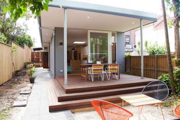 Rear exterior view of a compact contemporary home extension with a flat roof, covered outdoor dining deck, and stepped timber decking. Large glazed doors connect the deck to an open-plan interior, while a paved side path, timber boundary fencing, and established planting frame the space. Bright, casual indoor-outdoor living setting.
