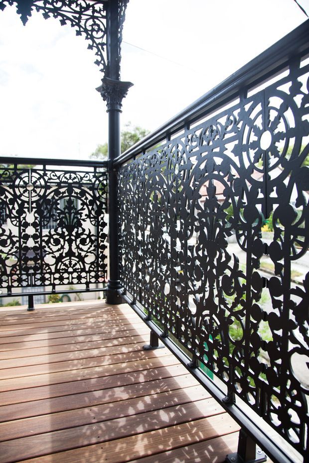 A close-up view of an elevated timber deck or balcony with ornate black metal privacy screens and balustrades. The screens feature intricate floral and scrollwork cut-out patterns, supported by decorative metal posts and rails. Bright daylight creates patterned shadows across the timber decking, with a suburban exterior visible beyond.