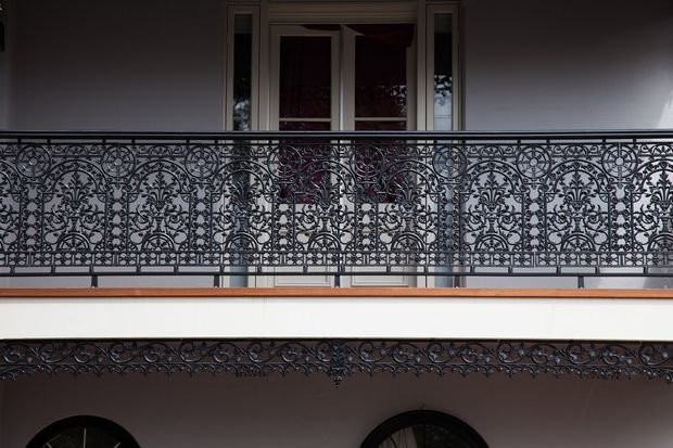 Close-up architectural view of an ornate cast-iron balcony or gallery railing with intricate floral and scrollwork patterns. A dark-framed opening and upper-level facade are visible behind it, with a matching decorative iron frieze beneath the balcony. The composition emphasizes heritage-style metalwork and layered architectural detailing.