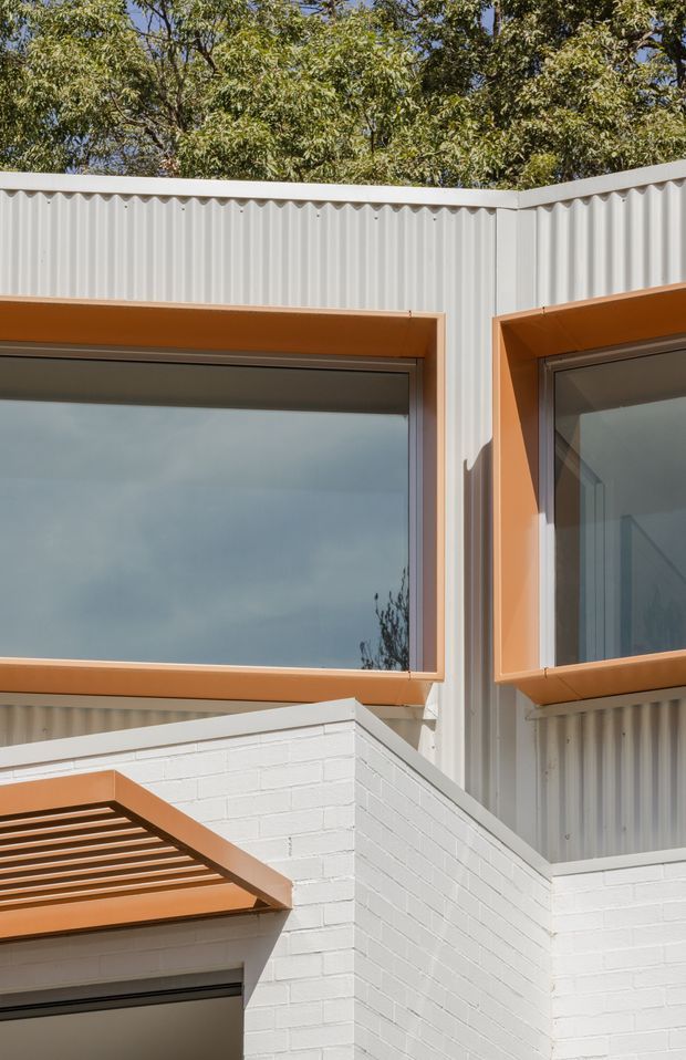 Close-up exterior architectural detail of a contemporary building, featuring white vertically profiled metal cladding, white painted brickwork, large dark glazed windows with orange-brown metal frames, and a matching horizontal louvre or pergola element. Trees are visible above the roofline.