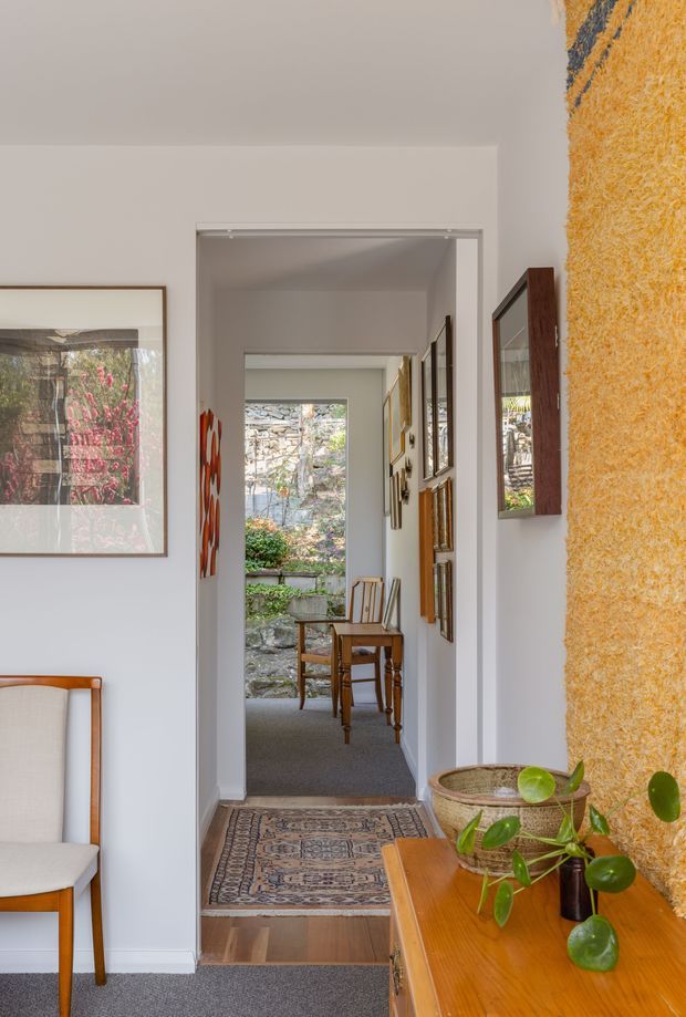 A bright residential hallway viewed toward a small sitting area and garden beyond. White walls and door trims frame a narrow corridor lined with framed artwork and mirrors, while a textured golden feature wall and warm timber console add character. A patterned runner, upholstered timber chair, potted plant, and glimpses of stone landscaping through the opening create a refined, eclectic interior.