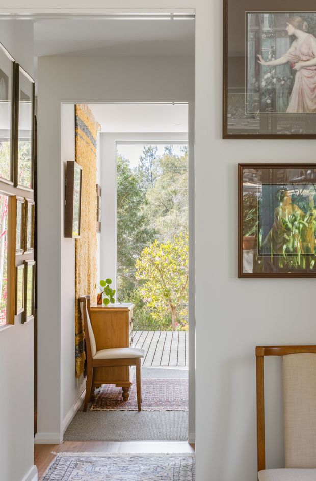 A bright, narrow residential hallway or entry passage leading toward an open doorway and timber deck. The space has white painted walls, framed artwork and mirrors, carpeted flooring with layered rugs, and a small timber console with a matching upholstered chair. A leafy garden is visible beyond the doorway. The styling is warm, curated, and traditional with natural timber accents.