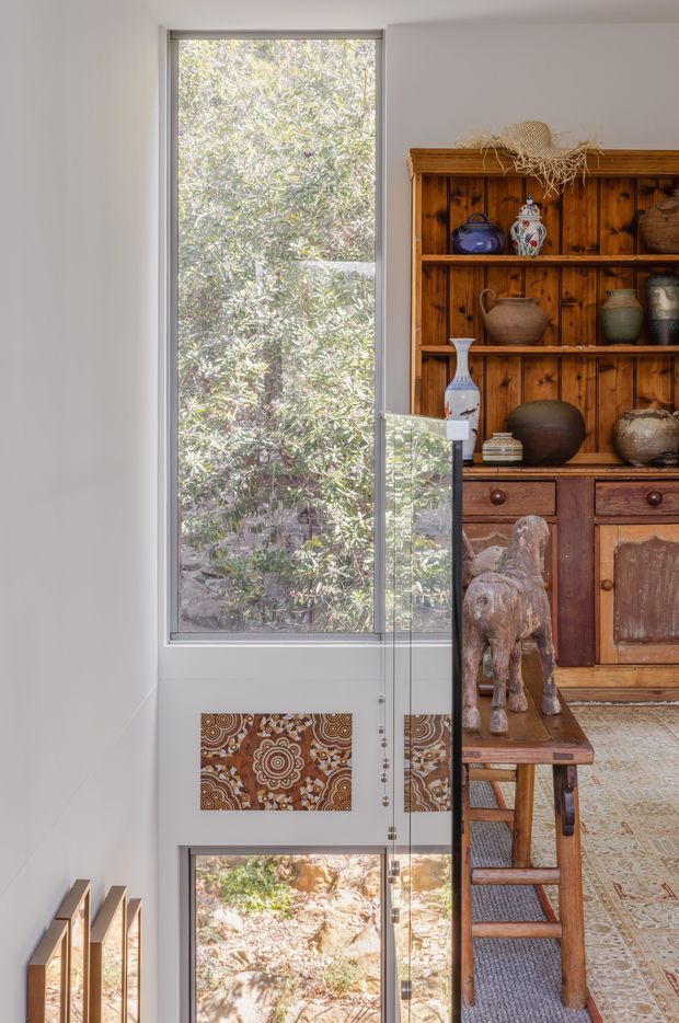 A bright, narrow interior stairway or landing with full-height glazing overlooking leafy vegetation. A timber display cabinet with open shelves, drawers, pottery, textiles and a carved animal sculpture occupies the right side, while a glazed stair balustrade and lower-level window are visible in the foreground. The space has warm rustic detailing contrasting with white walls and abundant natural light.