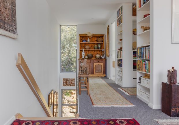 Bright upper-level hallway and landing overlooking a stairwell, with grey carpet, a long patterned runner, extensive white built-in bookshelves and storage, and a traditional wooden display cabinet at the far end. A tall window brings in views of greenery, while artwork and decorative objects add warmth.