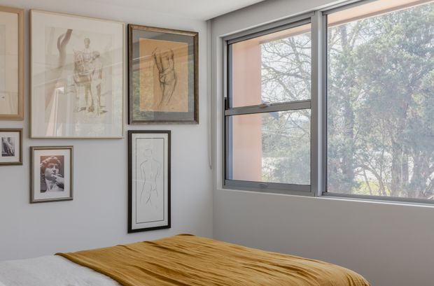 A compact, softly lit bedroom corner with a bed partially visible in the foreground, dressed in white bedding and a mustard-gold throw. A gallery wall of framed artwork occupies the left wall, while large aluminium-framed windows provide views of leafy trees outside. The space has a restrained, contemporary feel with pale grey walls and minimal furnishings.