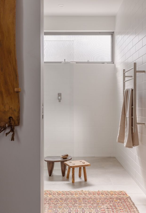 A bright, minimalist bathroom with white tiled walls, pale large-format floor tiles, a high horizontal frosted window, and a wall-mounted heated towel rail holding neutral towels. A timber door and patterned rug are visible at the entrance, while small wooden stools add warmth to the otherwise restrained space.