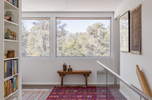 Bright, minimalist upstairs landing or hallway with large timber-framed windows overlooking trees, white walls and ceiling, a built-in white bookshelf, a slim timber console table, colourful patterned rugs, wall art, and a glass stair balustrade. The space has a calm, curated feel and receives abundant natural light.