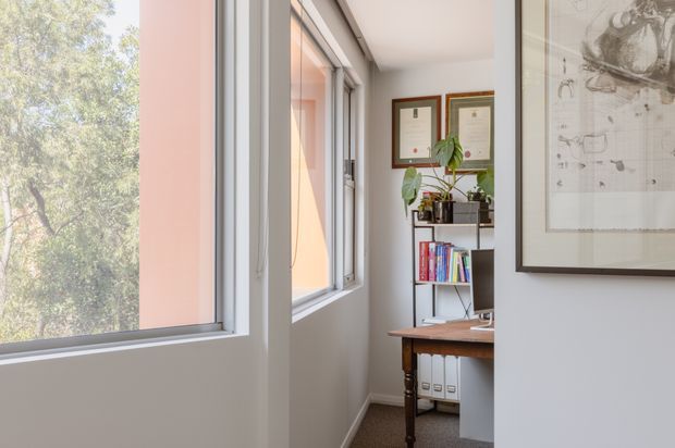 A bright, narrow home office or study viewed along a glazed corridor. Large white-framed windows overlook leafy trees, while a compact wooden desk, open shelving with books and files, framed certificates, and a potted plant create a functional workspace. The walls and ceiling are white, with warm daylight and a framed monochrome artwork adding character.