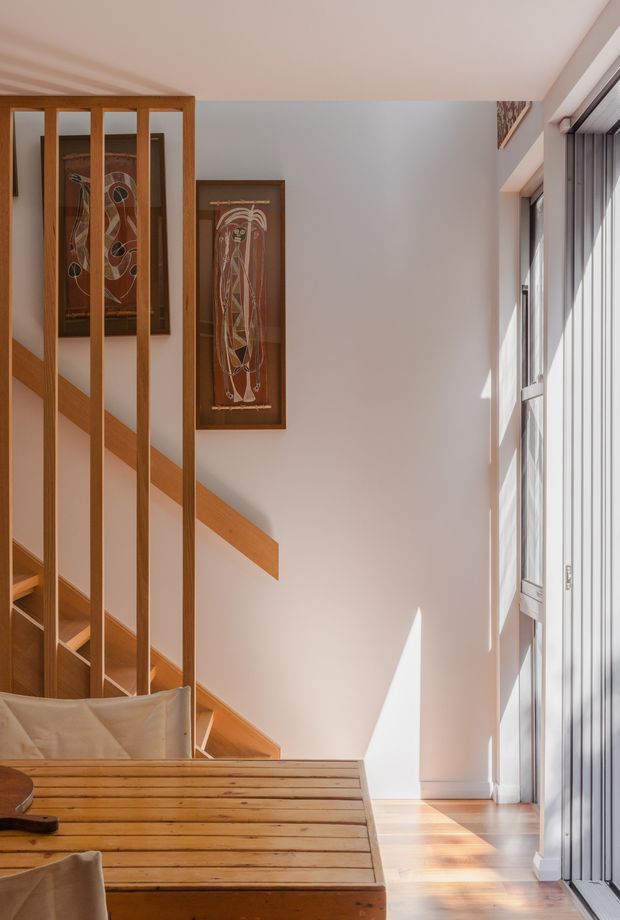 A bright, double-height interior stair hall with warm timber stairs, balustrade and slatted divider, pale plaster walls, tall glazed windows, and framed artworks. Sunlight creates strong geometric highlights across the timber floor and wall. A partial upholstered seating area is visible in the foreground.