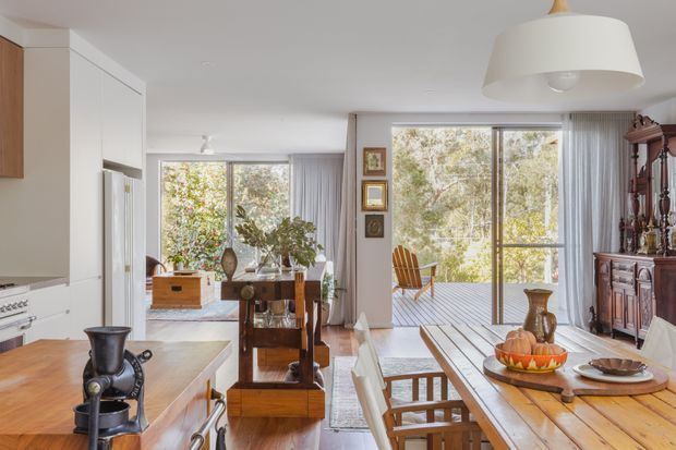 Bright open-plan interior combining a contemporary white kitchen with a timber dining area and living space. Large glazed doors and windows open to a covered deck, bringing in abundant natural light. Warm timber flooring, a substantial timber dining table, dark traditional sideboard, framed wall art, and vintage-style furnishings create a relaxed, eclectic character.
