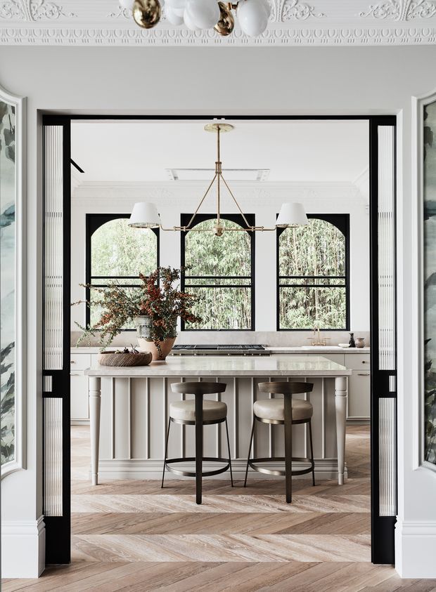 A refined, light-filled kitchen viewed through a wide black-framed opening. The room features a large pale island with fluted panel detailing and a light stone benchtop, two upholstered metal bar stools, black-framed arched windows, brass lighting, white cabinetry, ornate ceiling mouldings, and warm herringbone timber flooring. The styling combines classic architectural details with contemporary furnishings and finishes.