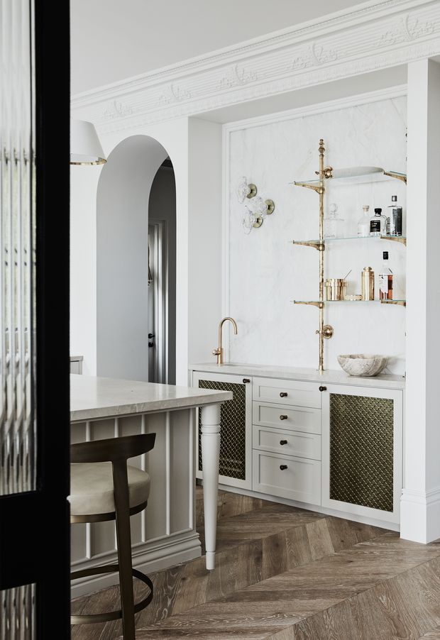 Elegant, light-filled kitchen with white classical mouldings, decorative cornice, arched doorway, marble-look splashback and benchtops, brass open shelving and tapware, and detailed white cabinetry with diamond-patterned metal grille panels. A peninsula with a dark timber bar stool occupies the foreground, while herringbone timber flooring adds warmth and texture.