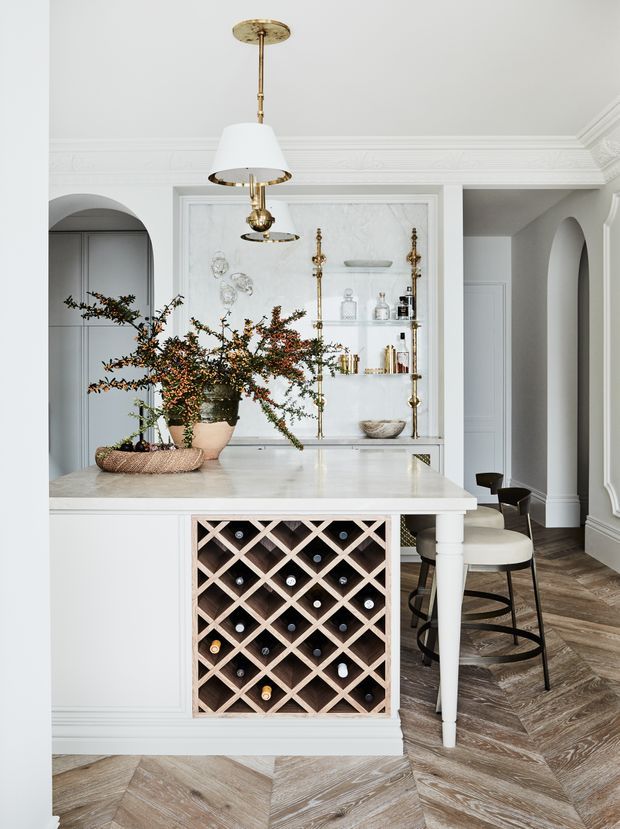 Elegant, light-toned kitchen or entertaining area featuring a marble-topped island with integrated diamond-pattern wine storage, brass-accented open shelving, decorative wall moulding, arched openings, pendant lighting, and herringbone timber flooring. Bar stools and a floral arrangement add warmth to the refined interior.