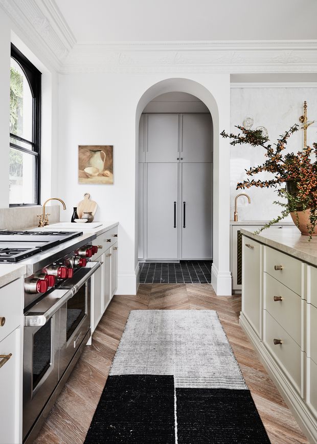 A bright, refined galley kitchen with white cabinetry, a large professional stainless-steel range, pale stone benchtops, black-framed windows, and a central island. The room features an arched opening leading to a tall pantry or storage area, ornate ceiling cornices and wall mouldings, brass-toned tapware and hardware, and herringbone timber flooring layered with black-and-white runners. A large floral arrangement and artwork add decorative warmth.