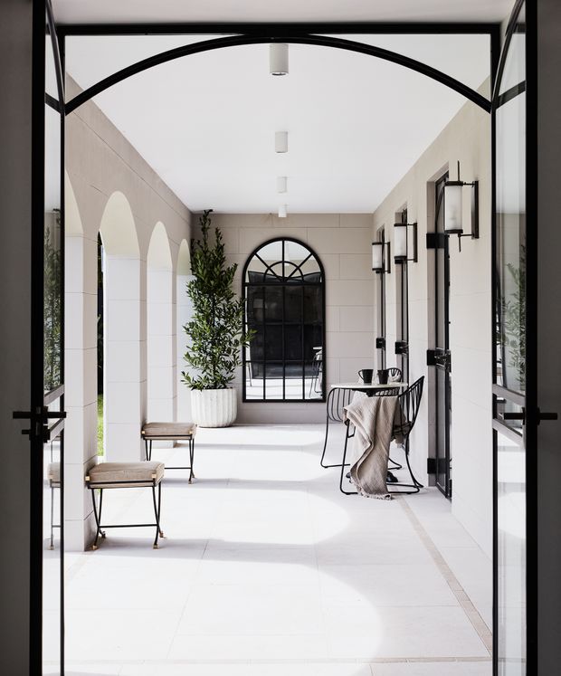 Bright, elegant enclosed courtyard or covered outdoor passage viewed through open black-framed glass doors. The space features pale tiled flooring, cream stone or rendered walls with arched openings, black steel-framed arched windows and doors, wall-mounted lanterns, a potted tree, and a small café-style table with chairs. The design has a refined contemporary Mediterranean influence and strong indoor–outdoor connection.