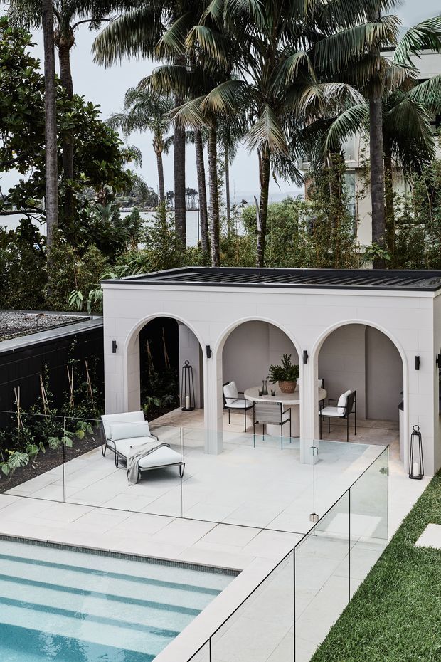 A refined outdoor pool terrace with a white arched pavilion featuring a covered dining and lounge area. Large pale stone or porcelain paving surrounds the pool, with frameless glass pool fencing, sculptural outdoor furniture, and lush tropical palm landscaping. The pavilion has a dark low-pitched roof and crisp white rendered walls, creating a resort-like contemporary setting.