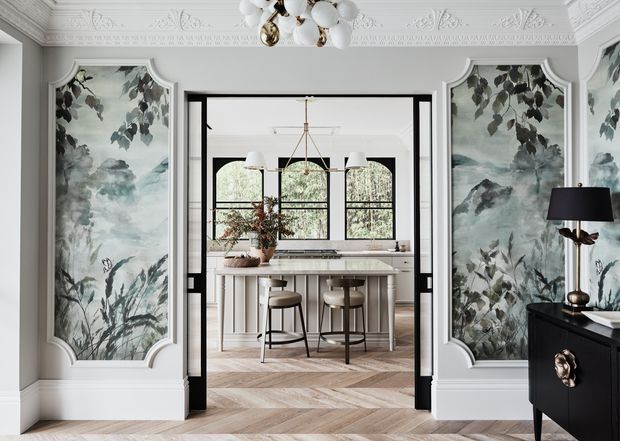 Elegant traditional interior view through open black-framed double doors into a bright kitchen or dining area. The foreground room has pale grey walls, ornate white ceiling cornices and decorative moulding, large framed botanical mural panels, and a black console with sculptural brass hardware. Beyond is a light-toned kitchen or dining space with a central island, stools, pendant lighting, arched black-framed windows, and herringbone timber flooring. The overall look combines refined heritage detailing with contemporary furnishings.