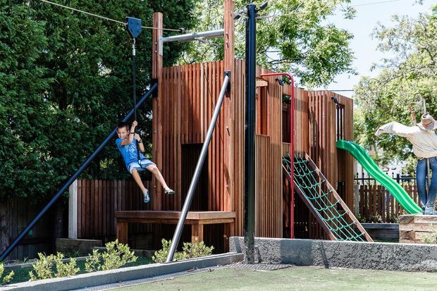 Outdoor children's playground in a landscaped setting, featuring a timber-clad play structure with climbing nets, swings, a green slide, and safety surfacing. Mature trees and planting surround the play area.